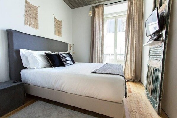 Un dormitorio con una cama, una mesita de noche, un televisor montado en la pared y un ventanal con cortinas.