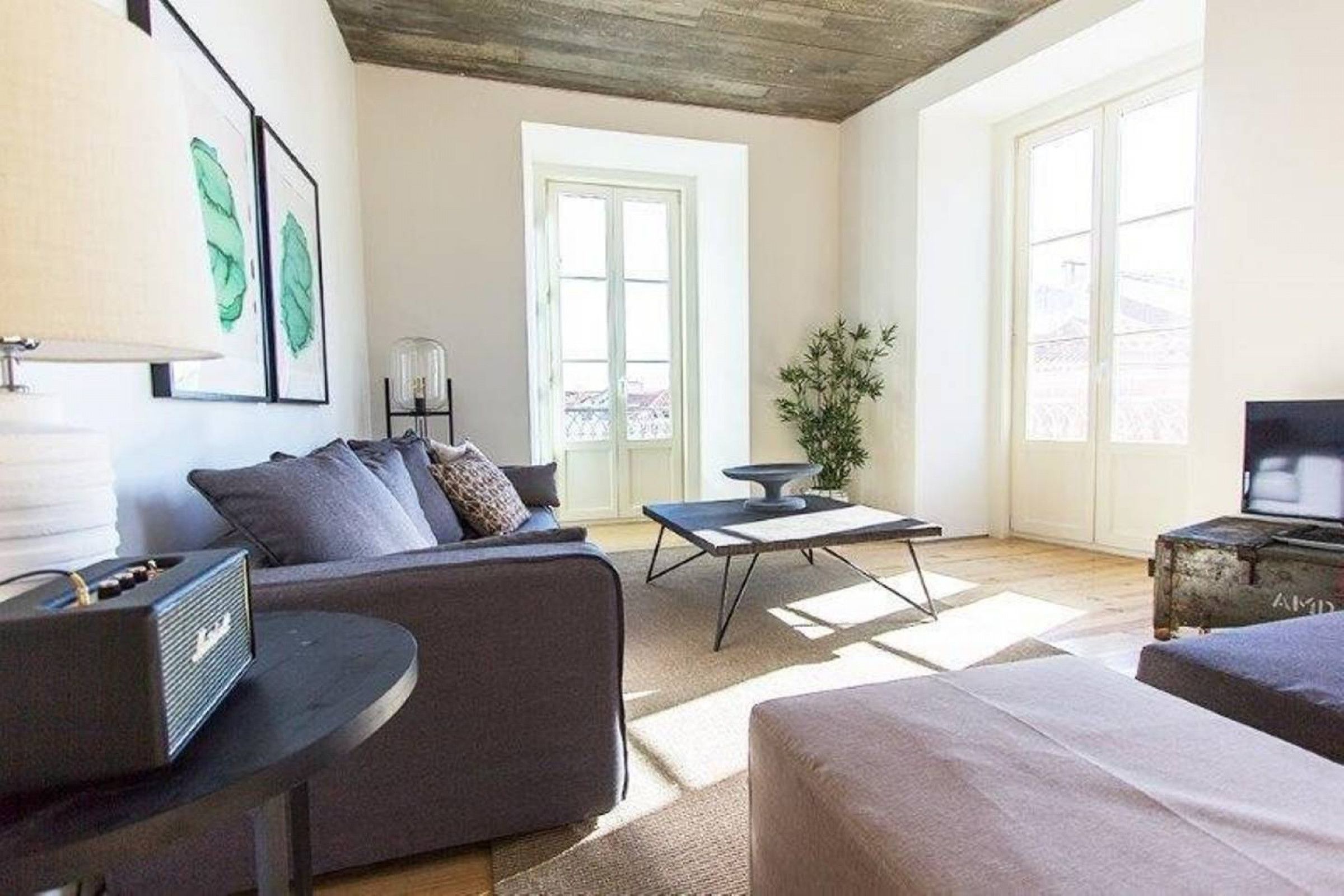 A modern living room with a grey sofa, a coffee table, and a television. There are large windows allowing natural light and a view of the outside.