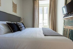 Una cama bien hecha con edredón blanco y almohadas estampadas, un televisor sobre un soporte de madera, y una ventana con cortinas.