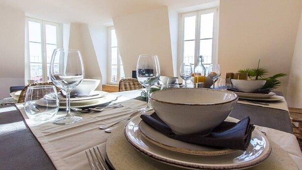 A dining table set with plates, bowls, and wine glasses, with a view of a balcony through the window.
