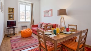 A cozy living room with a wooden dining table, orange chairs, a red sofa, and a television on a small table.