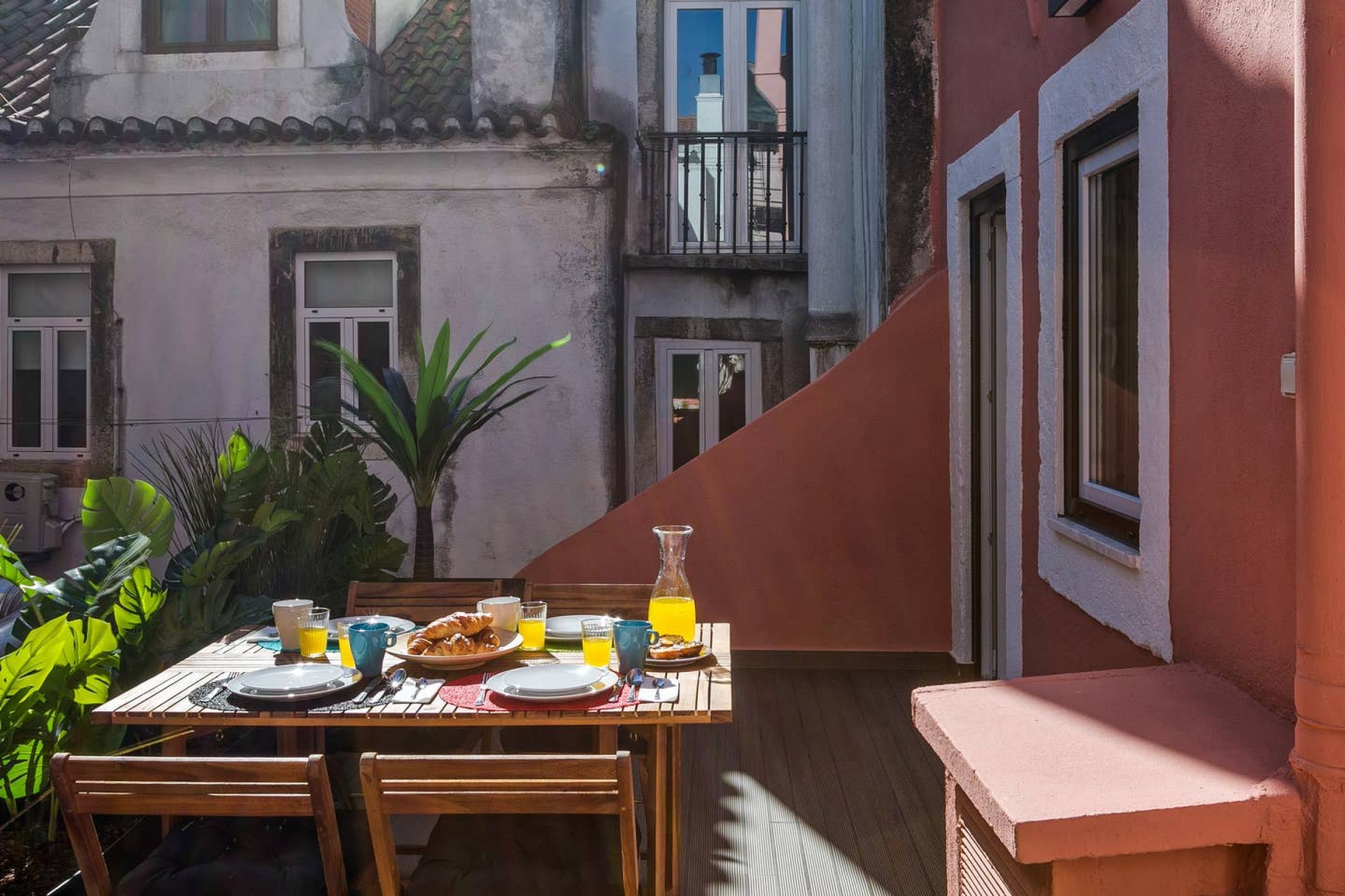 Un espace de restauration en plein air avec une table dressée pour le petit-déjeuner, entouré de plantes et bordé d’un bâtiment doté de balcons.