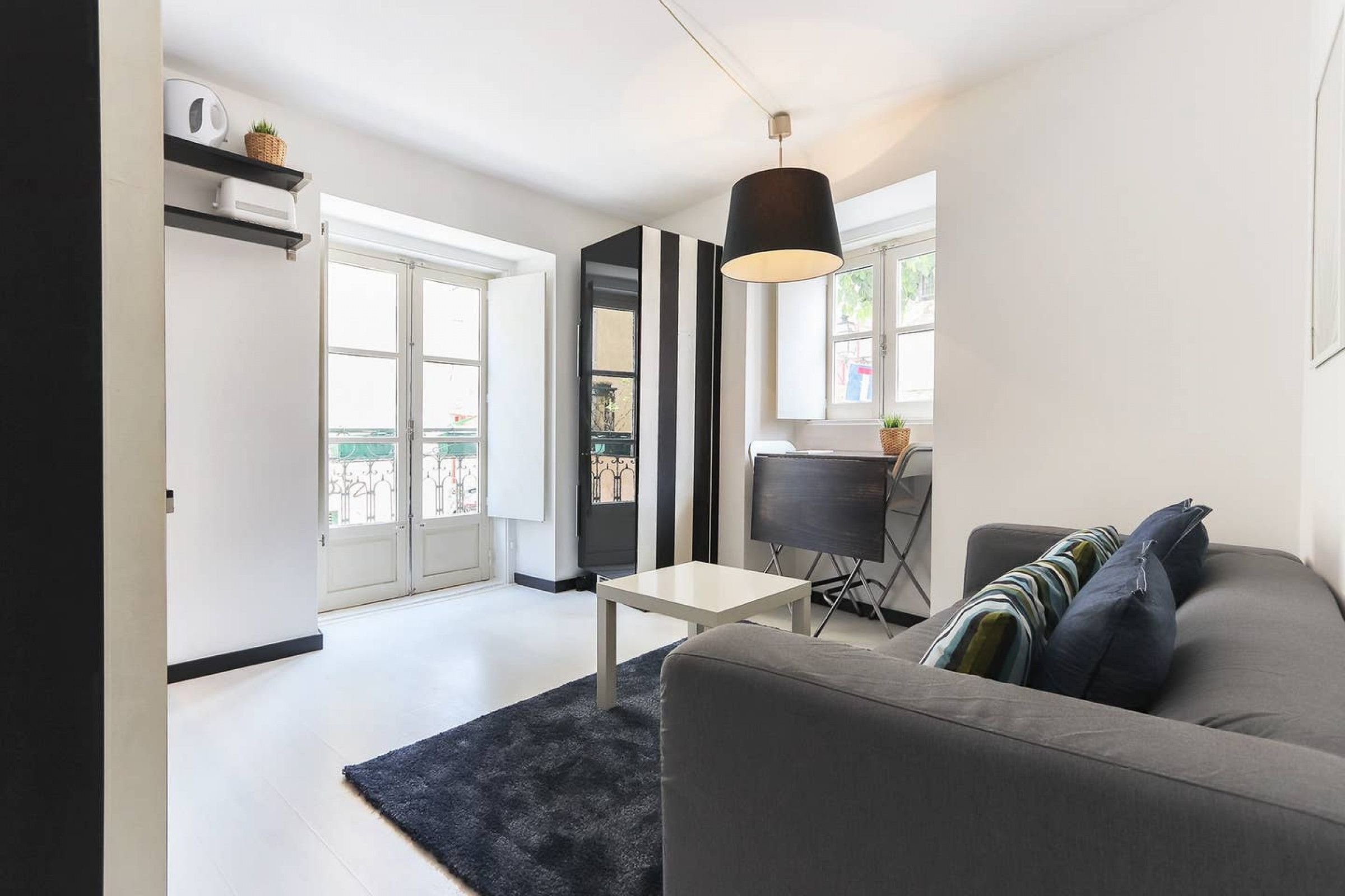A modern living room with a grey sofa, a small white table, and a black lamp. There is a door leading to a balcony with a view of the city.