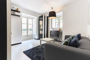 A modern living room with a grey sofa, a small white table, and a black lamp. There is a door leading to a balcony with a view of the city. - Atalaia · Tailor Made Flat in Central Bairro Alto (Lisboa)