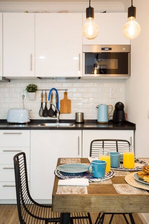 A modern kitchen with white cabinets, a stainless steel oven, and a dining area with a wooden table set for breakfast. - Metade · Charming Studio in Lisbon's Center (Lisboa)