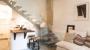 Una cocina moderna con una escalera de vidrio, un comedor con taburetes y una sala de estar con sofá y cojines.