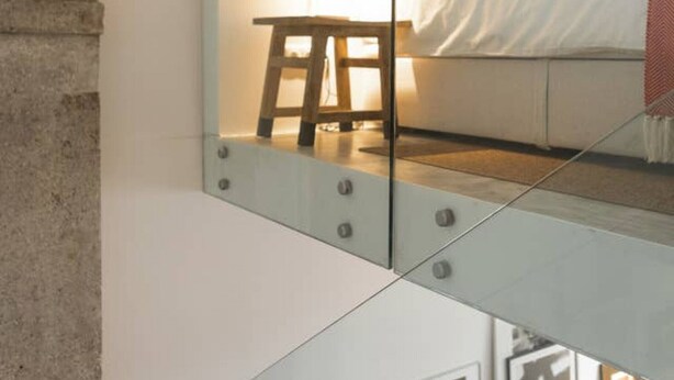 Un dormitorio moderno con separador de vidrio, cama con ropa de cama blanca, mesita de noche con lámpara y cuadros en la pared.