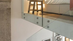 Un dormitorio moderno con separador de vidrio, cama con ropa de cama blanca, mesita de noche con lámpara y cuadros en la pared.