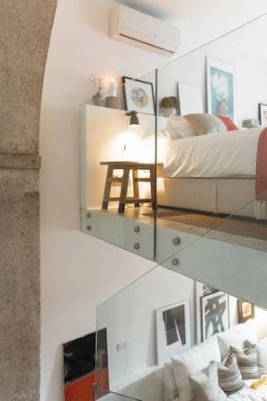 Un dormitorio moderno con separador de vidrio, cama con ropa de cama blanca, mesita de noche con lámpara y cuadros en la pared.