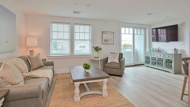 A living room with a sofa, a coffee table, and a chair. There is a TV mounted on the wall and a window with blinds. - Premier Ocean View Corner Condo (875S.F.) (Plymouth)