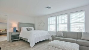 A modern bedroom with a bed, bedside table, lamp, and a grey sofa with an ottoman. - Premier Ocean View Corner Condo (875S.F.) (Plymouth)