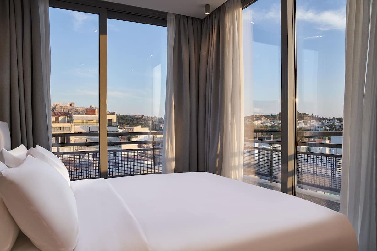 Una camera d'hotel con un letto grande, vista sulla città attraverso finestre a tutta parete e un balcone con ringhiera.