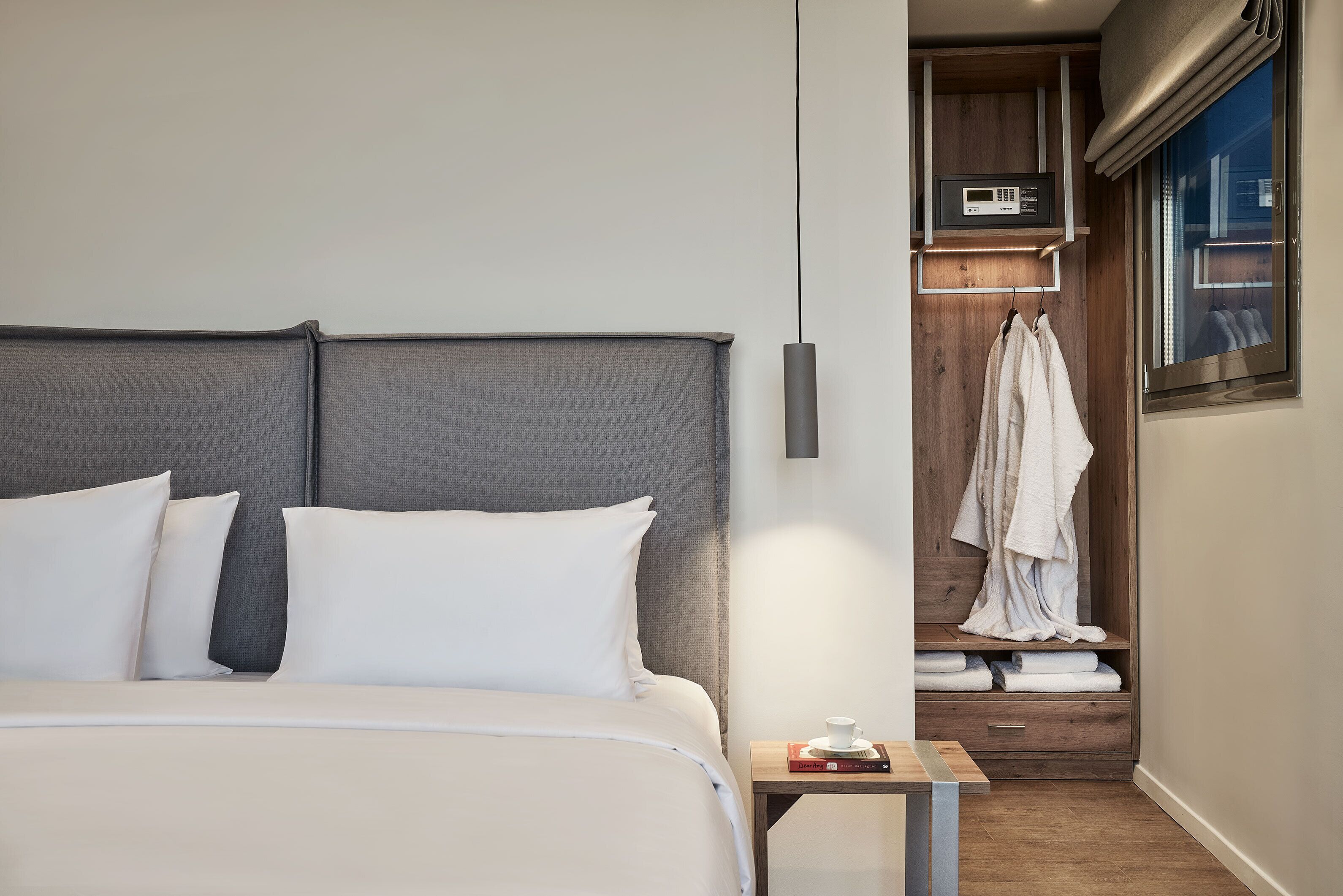 Una cama bien hecha con ropa blanca, una mesita de noche con una taza y un libro, y un armario de madera con albornoces colgados.