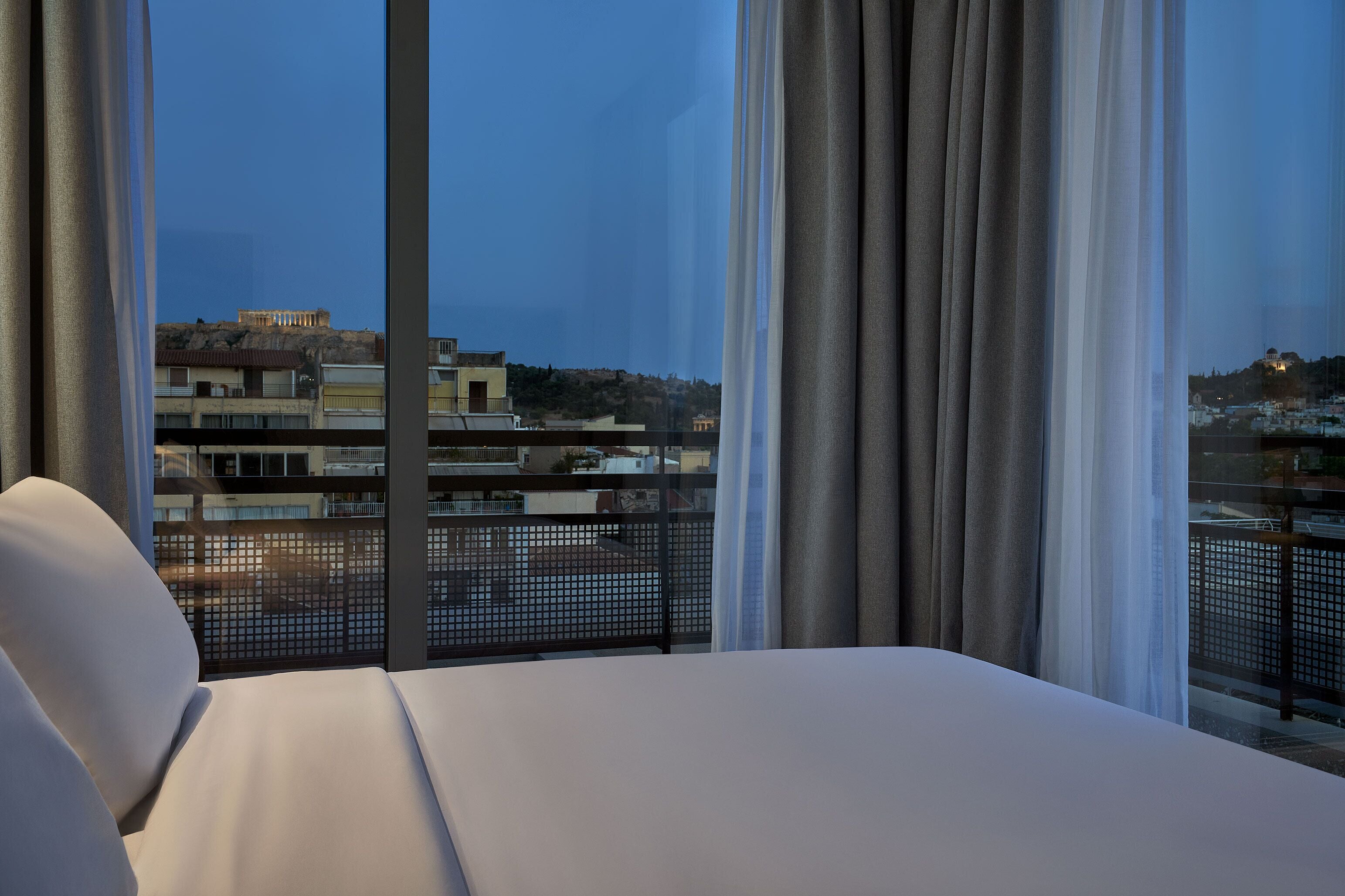 A hotel room with a large window, a bed, and a view of buildings and a hill.