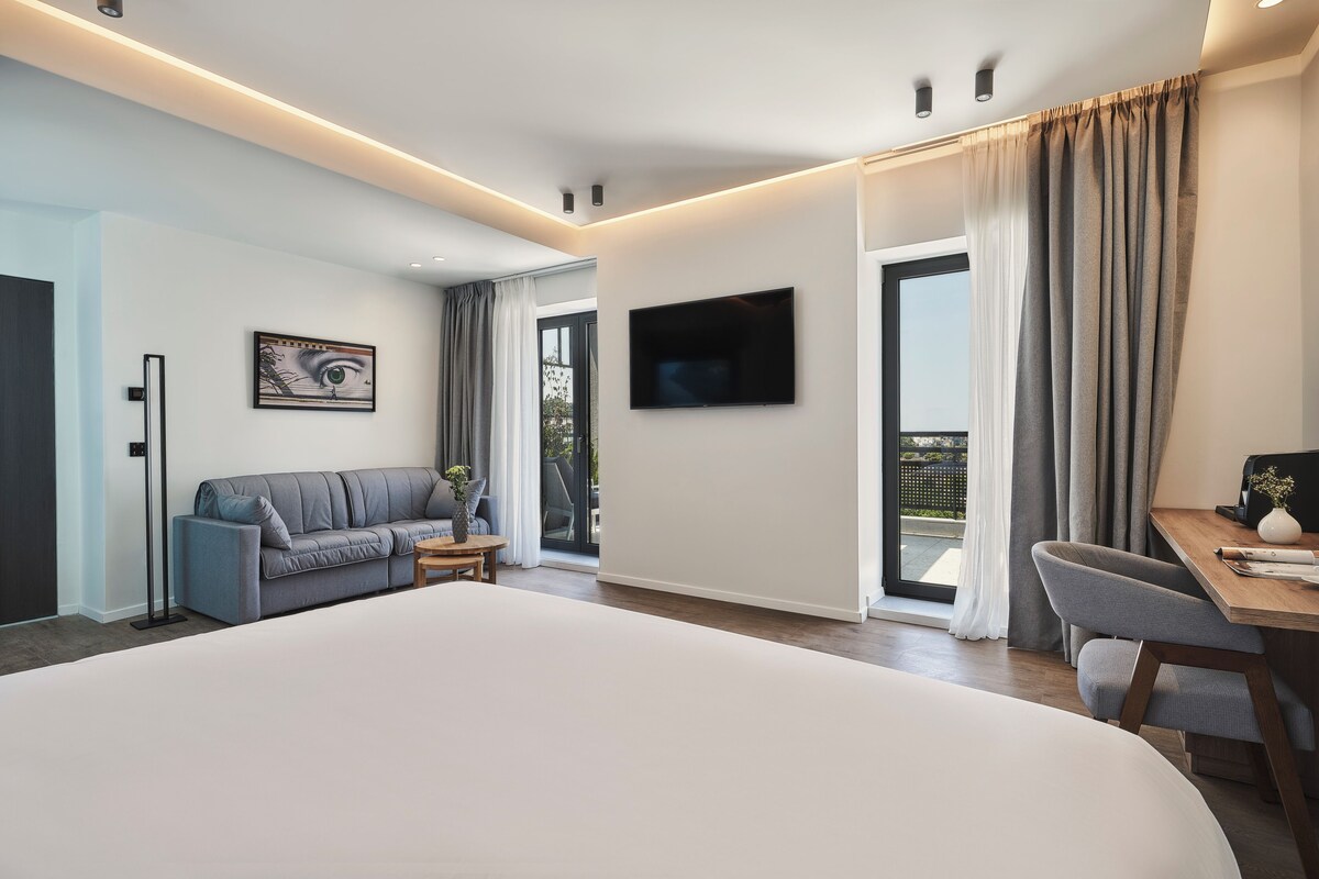 Habitació d'hotel moderna amb un llit gran, un sofà, un escriptori, una televisió i un balcó amb cortines.