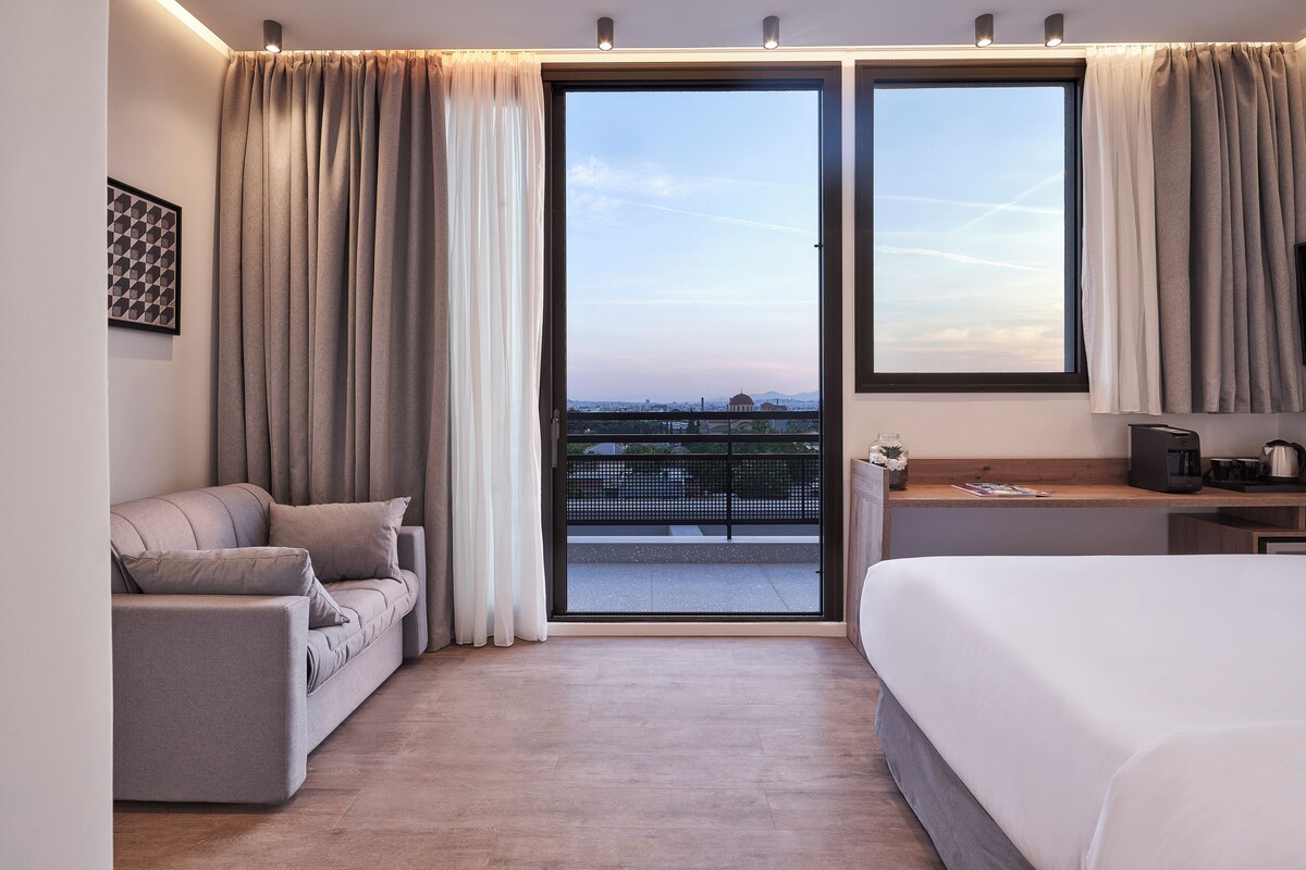 Habitació d'hotel amb llit, sofà, cafetera i vista a la ciutat a través de grans finestres.