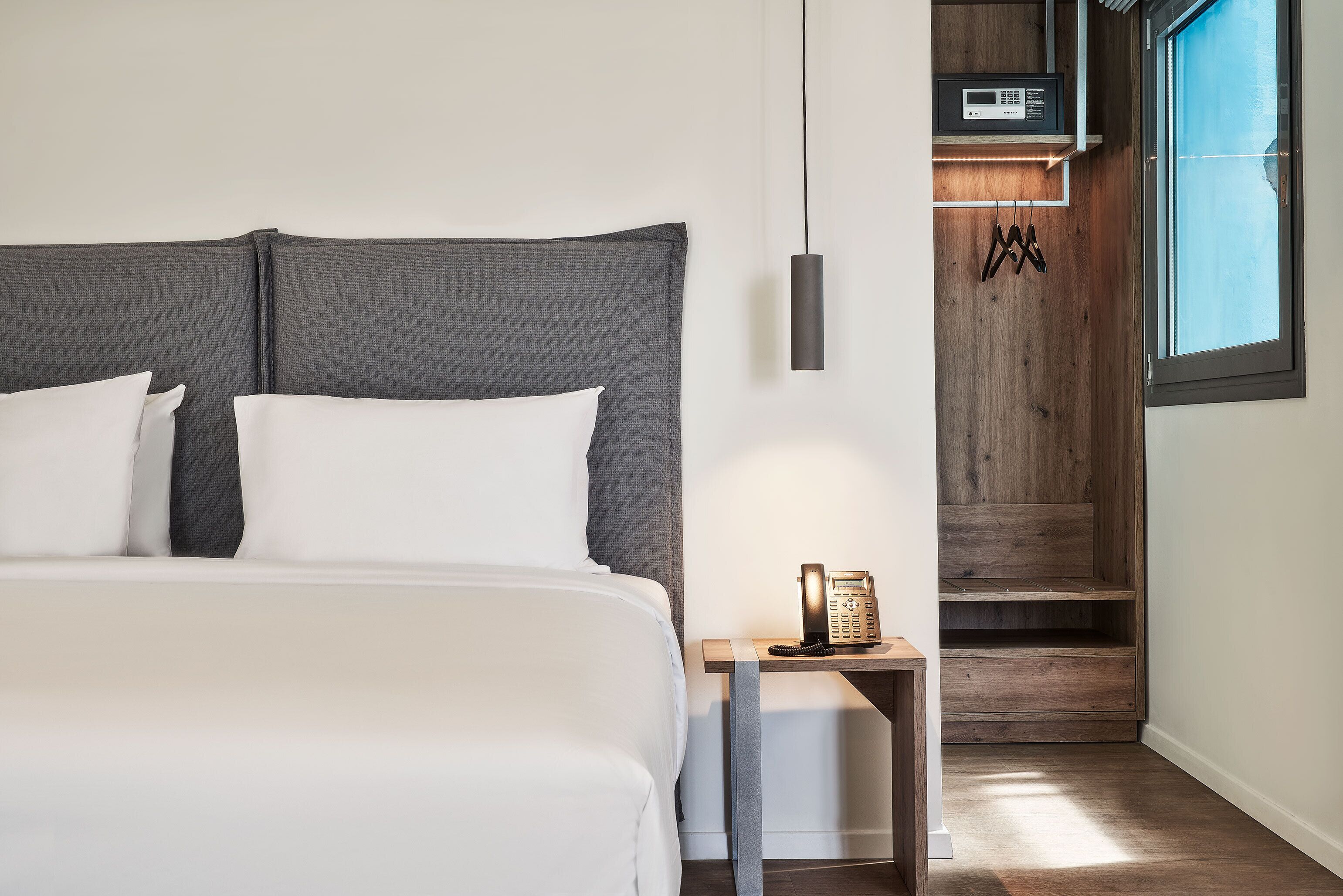 A modern hotel room with a large bed, a bedside table with a telephone, and a wooden shelving unit.