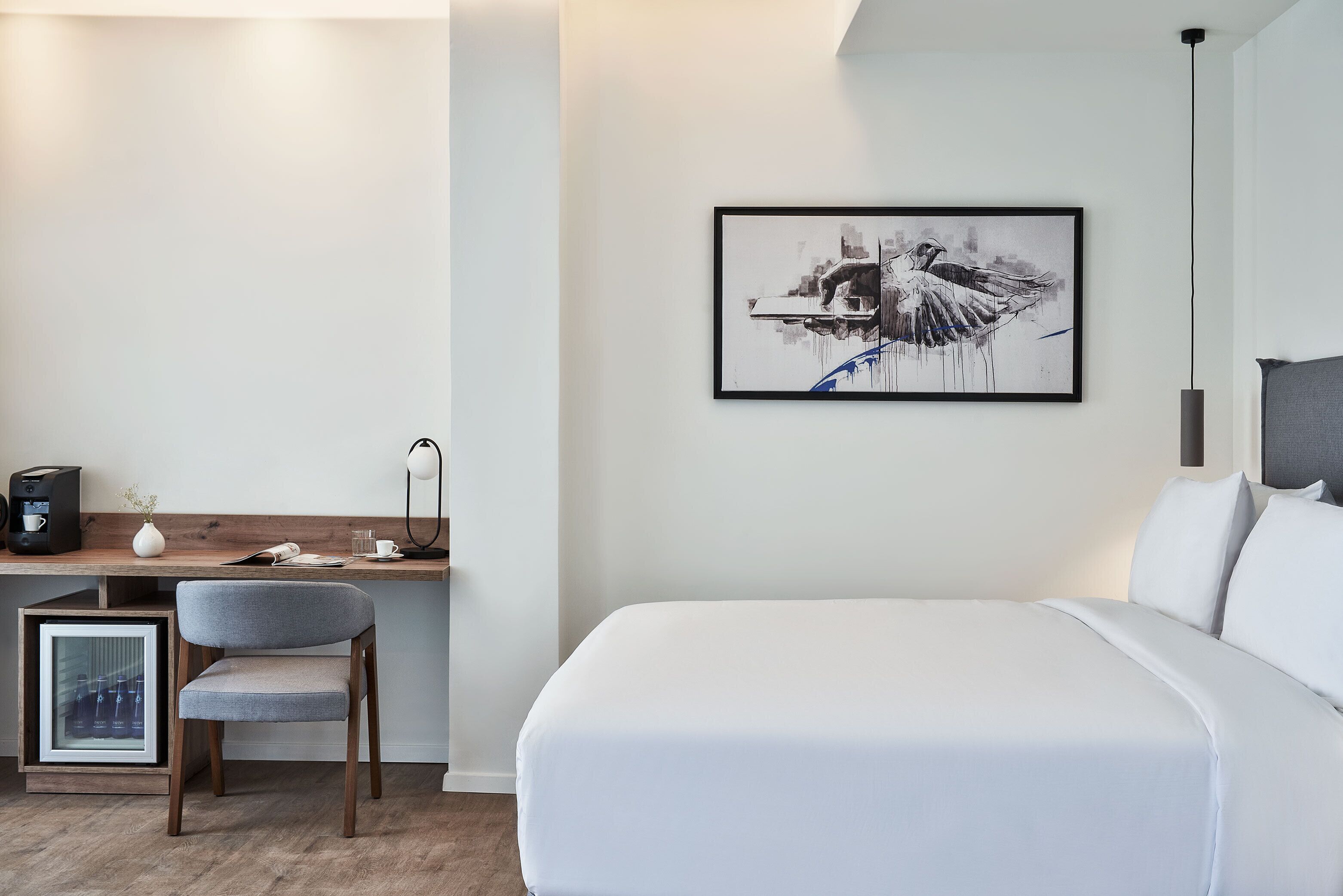 Une chambre d’hôtel moderne avec un lit soigneusement fait, un bureau avec une chaise, une œuvre d’art encadrée au mur et une machine à café.