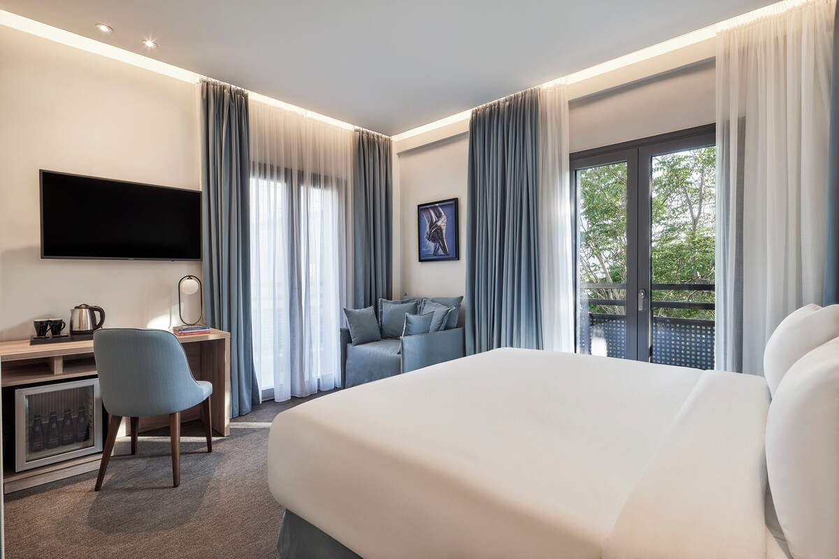 Habitació d'hotel amb llit, escriptori, cadira, televisió i balcó amb cortines.
