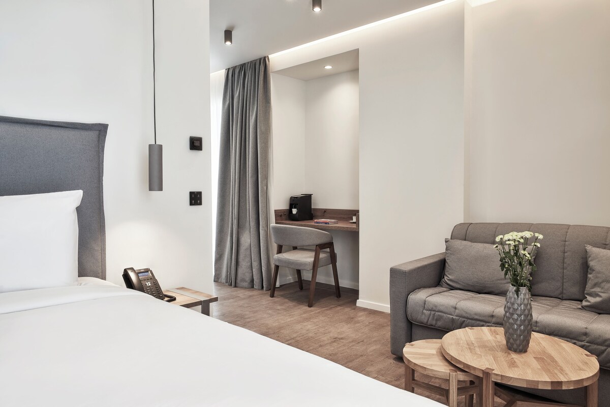 Habitació d'hotel moderna amb un llit, un escriptori, una cadira, un sofà i una taula rodona petita amb un ram.