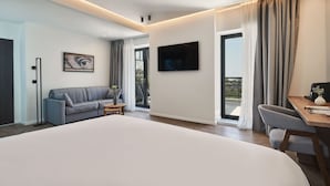 A modern hotel room with a large bed, a sofa, a desk, a TV, and a balcony with curtains. - NLH KERAMEIKOS - Neighborhood Lifestyle Hotels (Athens)