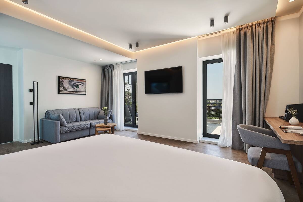 Una camera d'albergo moderna con un letto grande, un divano, una scrivania, una TV e un balcone con tende.