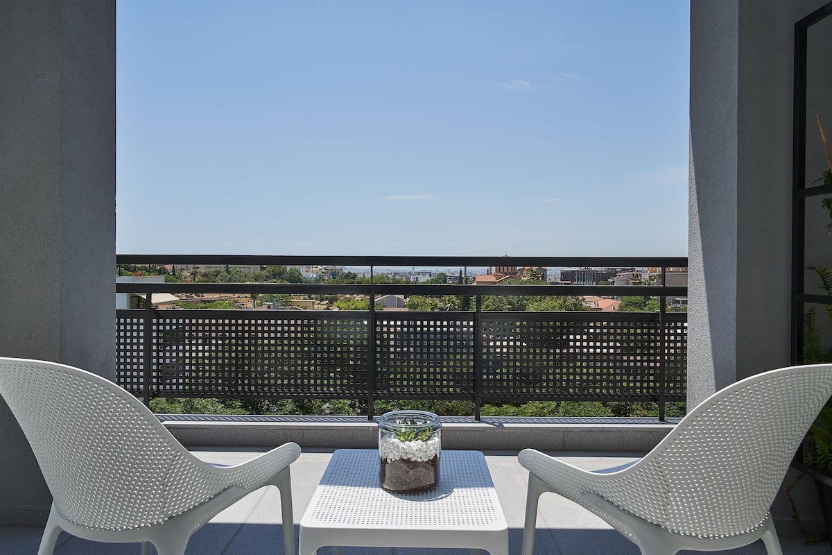 Un balcone con due sedie bianche, un vaso di vetro con fiori bianchi e vista sulla città.