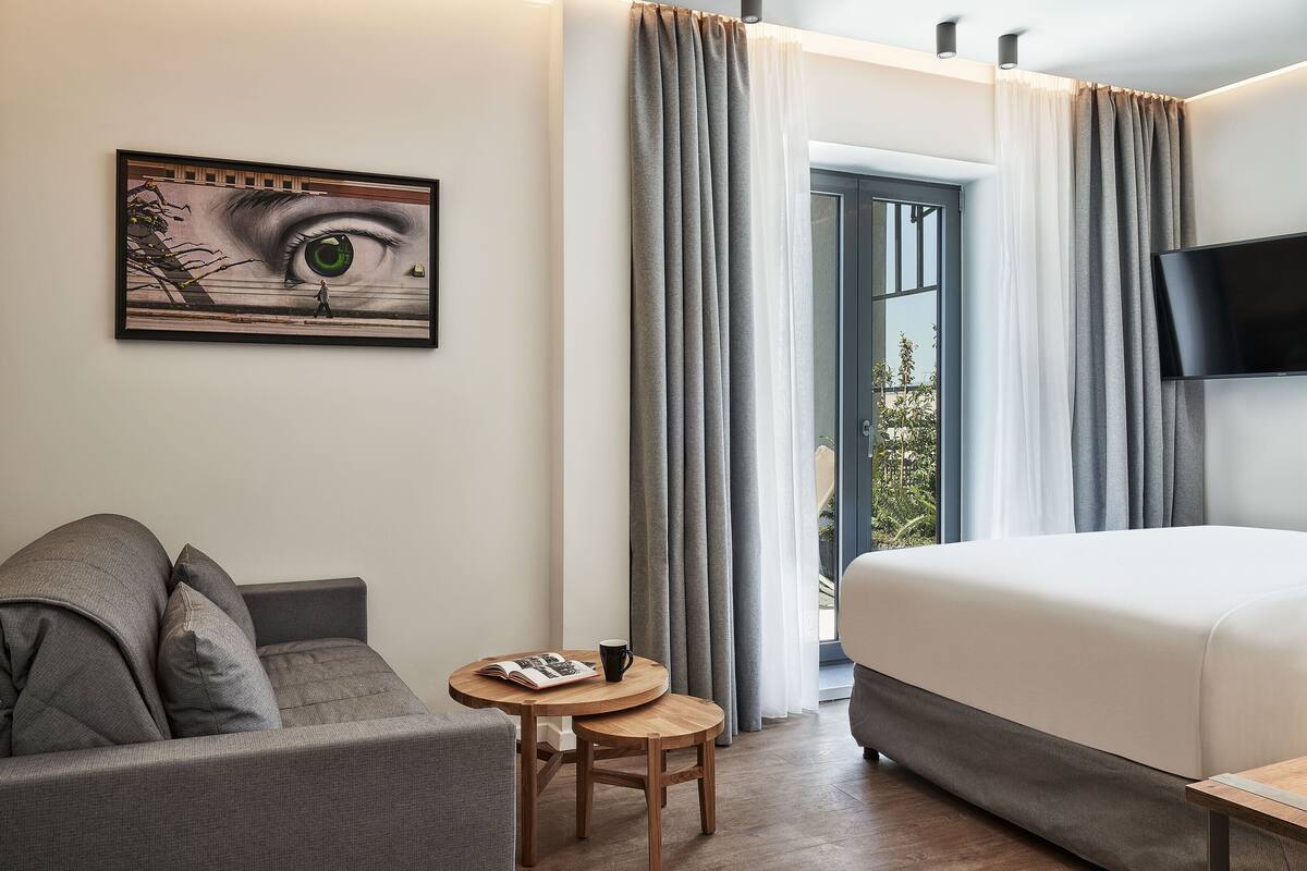 Camera d'albergo con un letto, un divano, un tavolino con una rivista e una tazza, un quadro raffigurante un occhio sulla parete e un televisore fissato al muro.