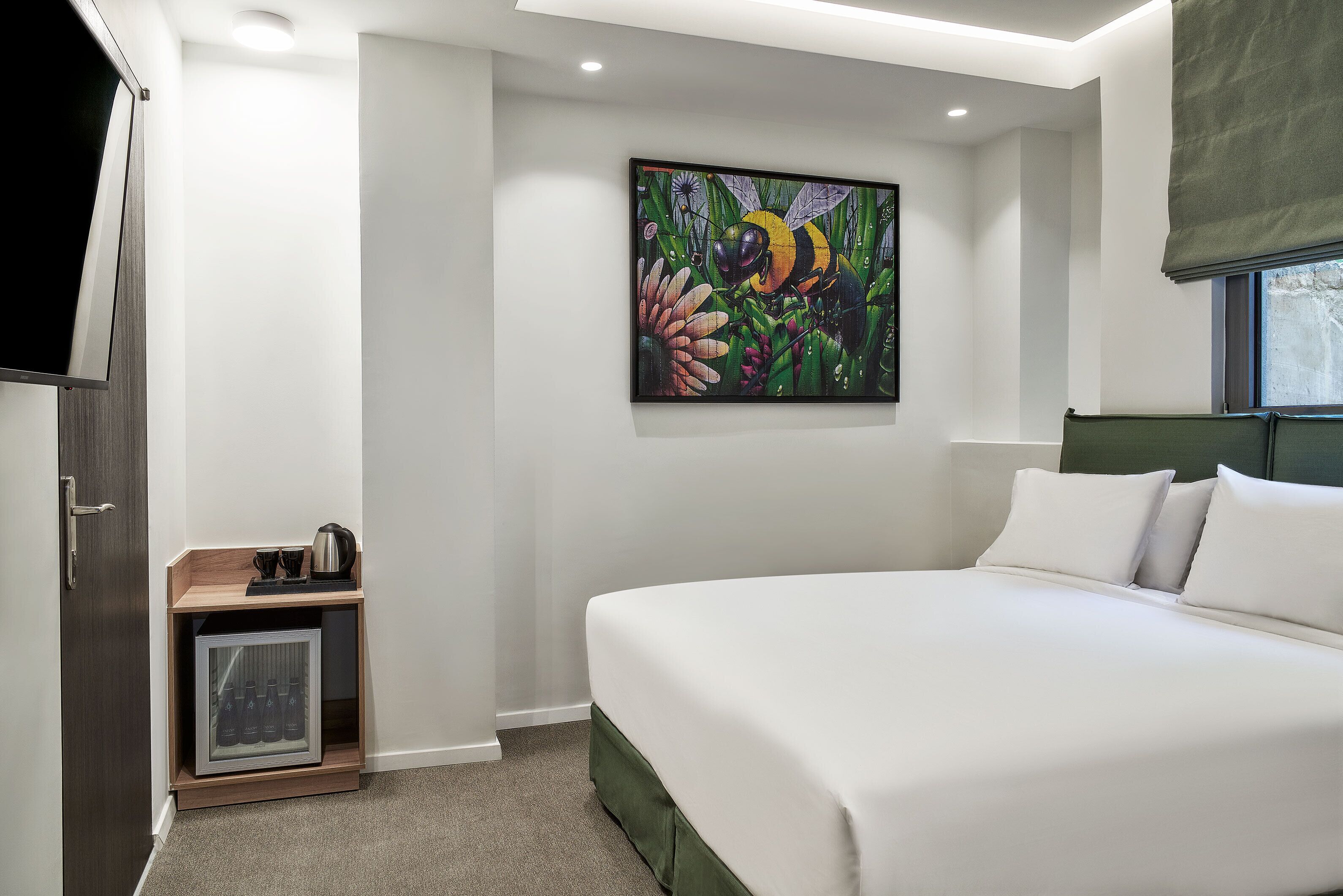 A bedroom with a large bed, a painting of a bee on the wall, a small cabinet with a kettle, and a window with a view.
