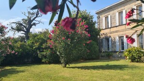 A well-maintained garden with a large tree and blooming flowers, a stone building with white shutters, and a clear blue sky. - Gîte Domaine de La Gravette (Cubnezais)