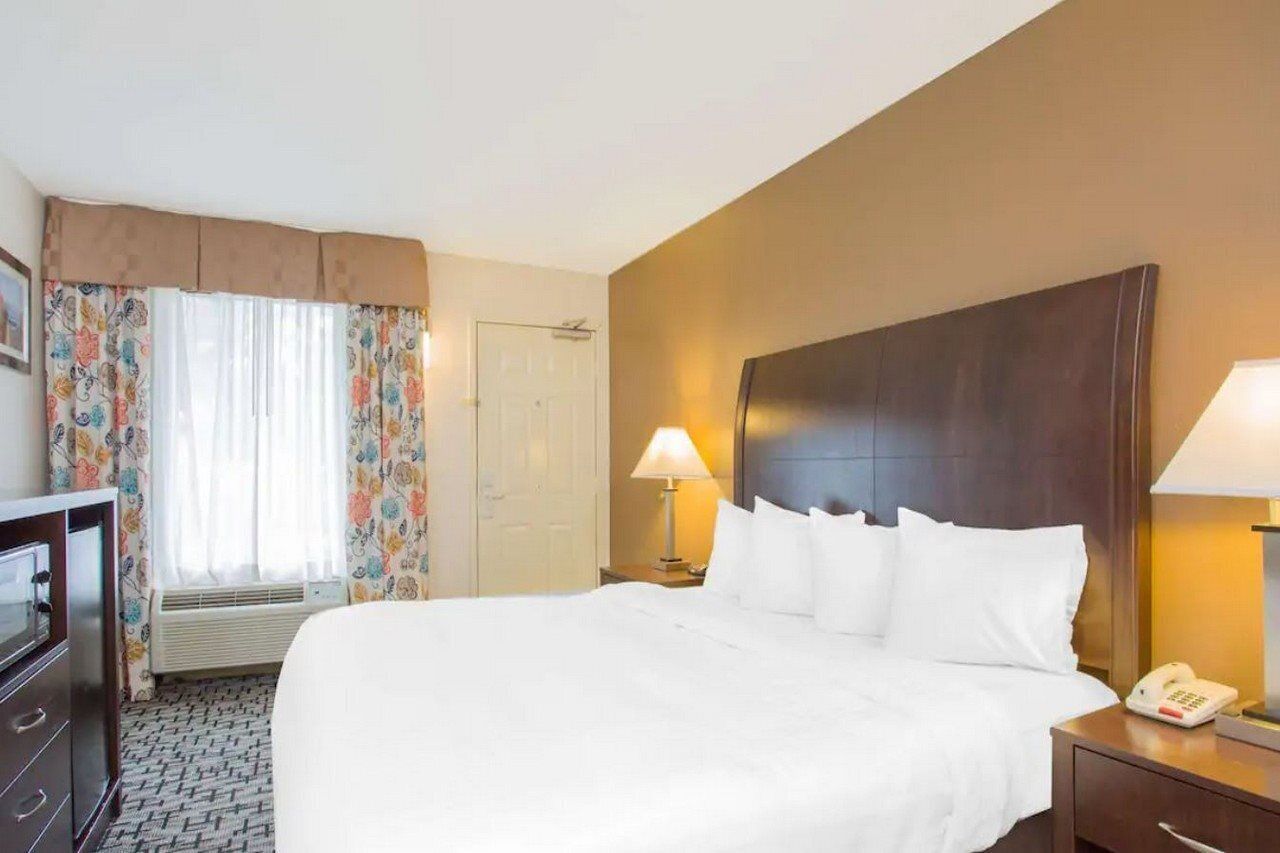 A hotel room with a large bed, two bedside lamps, a telephone, a desk with a lamp, a wardrobe, and a window with curtains.