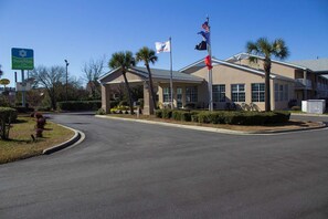 A hotel entrance with a curved driveway, palm trees, and flags. - Surestay Hotel by Best Western North Myrtle Beach (North Myrtle Beach)