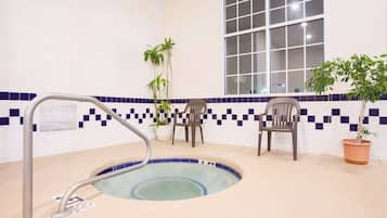A hot tub with a metal ladder, two chairs, and potted plants in a room with tiled walls and a window.