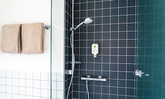 Un baño con mampara de vidrio, un perchero con dos toallas, un cabezal de ducha manual, un grifo empotrado en la pared y un dispensador de jabón.
