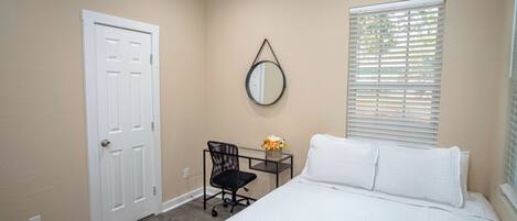 A bedroom with a bed, a desk, a chair, a door, and a window with blinds.