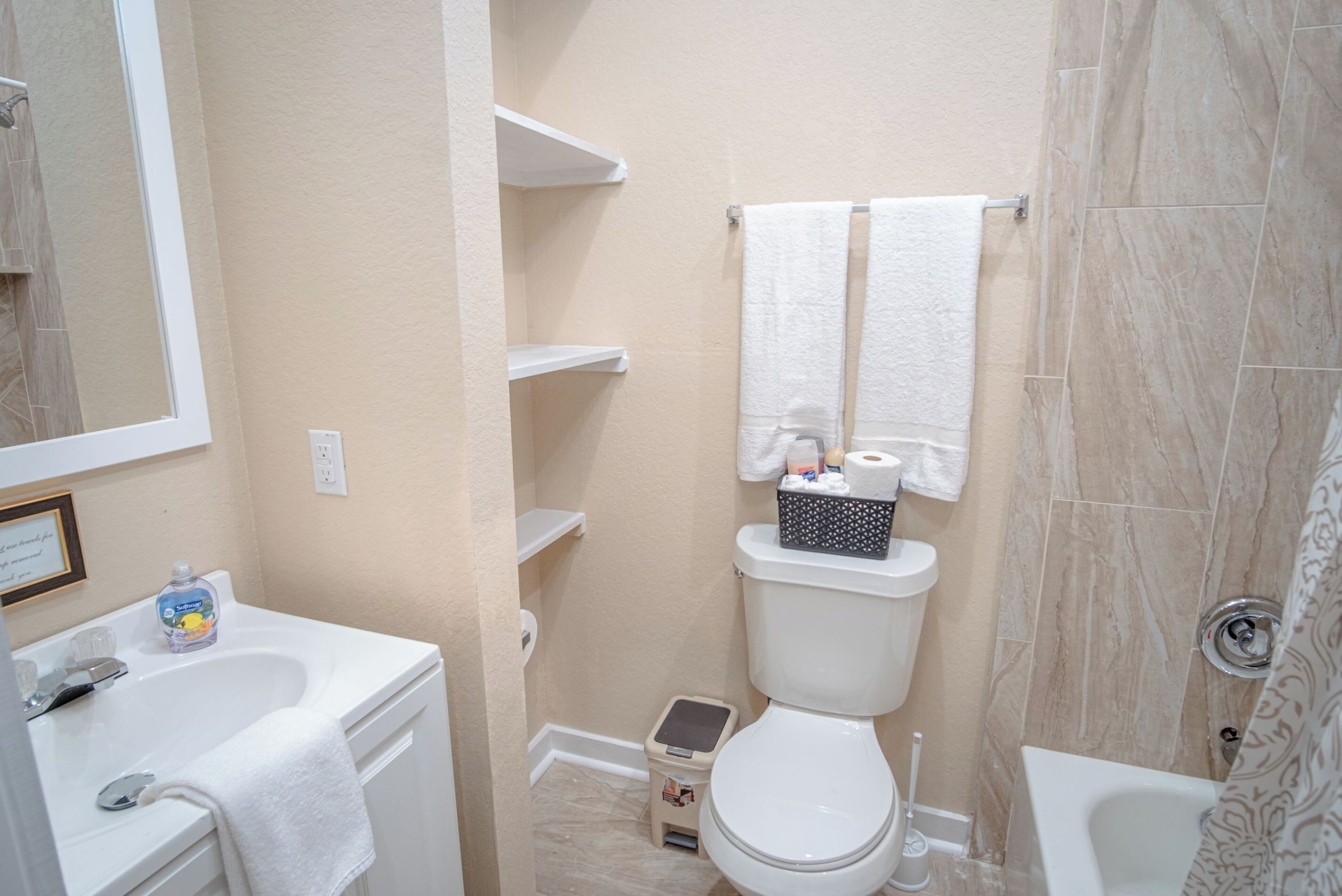 Un baño con inodoro, lavabo y ducha. Hay toallas blancas colgadas en la pared, un taburete pequeño y una botella de jabón de manos sobre el lavabo.