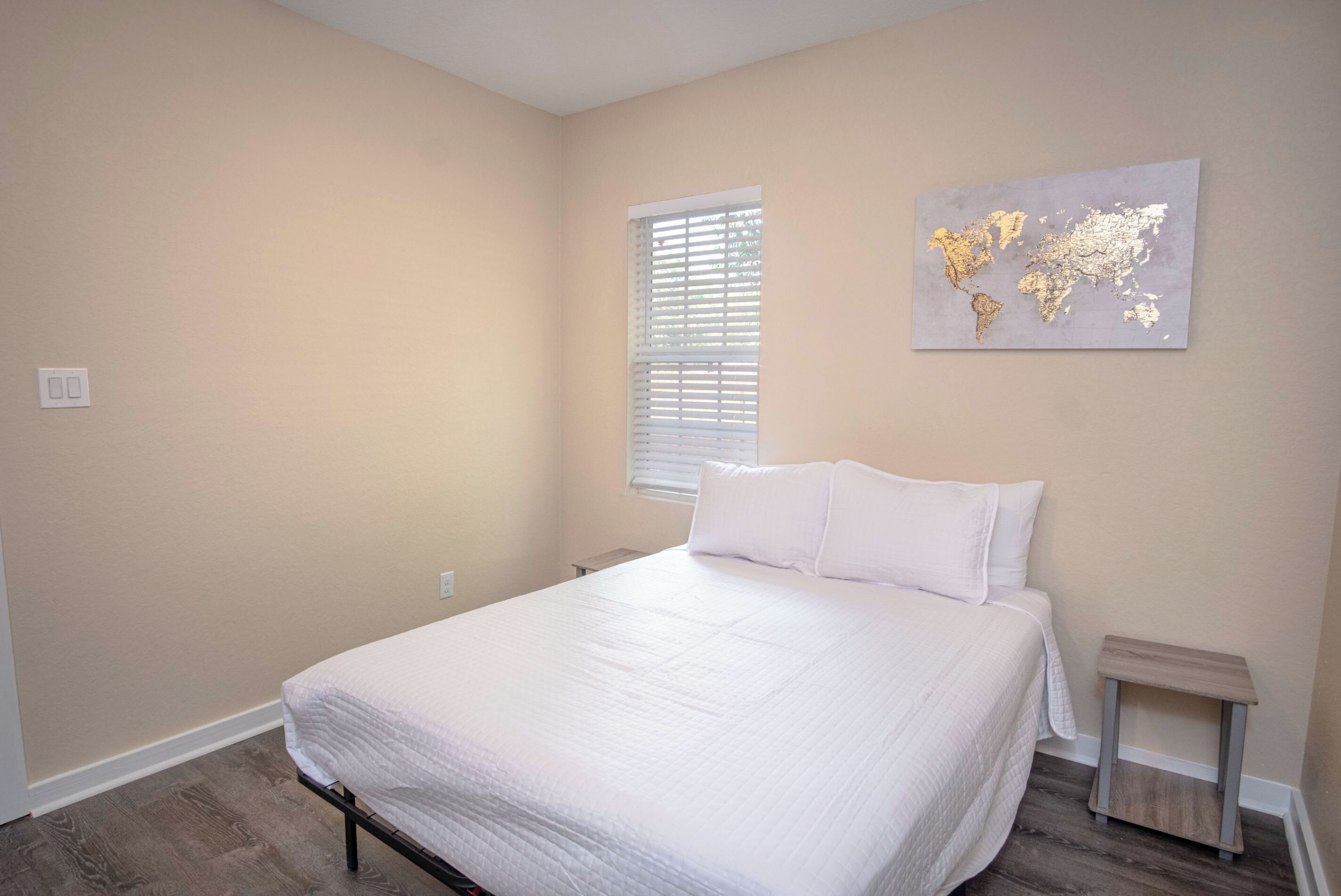 A bedroom with a bed, a window with blinds, a small bedside table, and a world map artwork on the wall.