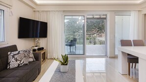 A modern living room with a flat-screen TV, a sofa with patterned cushions, a dining table with chairs, a potted plant, and a sliding glass door leading to a balcony with outdoor furniture. - Family-Friendly villa w/Pool, Children area & BBQ (Vlichada)