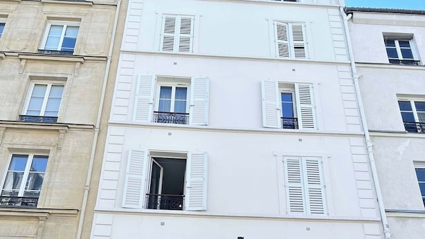 Un immeuble à plusieurs étages, doté de façades blanches et de volets, d’une marquise rouge et d’une devanture visible avec une enseigne indiquant « Restaurant ».