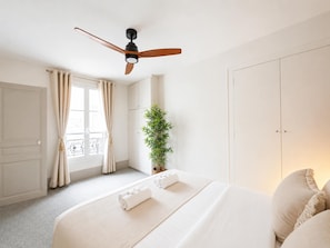 A bedroom with a bed, a ceiling fan, a window with curtains, a potted plant, and a wardrobe.