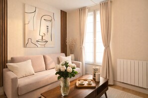 Uma sala de estar com um sofá, uma mesa de centro com um vaso de flores, um quadro na parede e uma janela com cortinas.