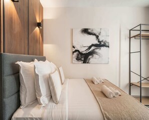 A modern bedroom with a grey headboard, white bedding, a beige throw blanket, and a black and white abstract painting on the wall.