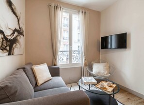 A living room with a sofa, a glass coffee table with a book and a tray, a chair, a TV, and a window with curtains.