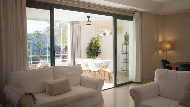 Ein modernes Wohnzimmer mit einer Couch, einem Esstisch mit Stühlen und Blick auf einen Balkon mit Pflanze und Tisch.
