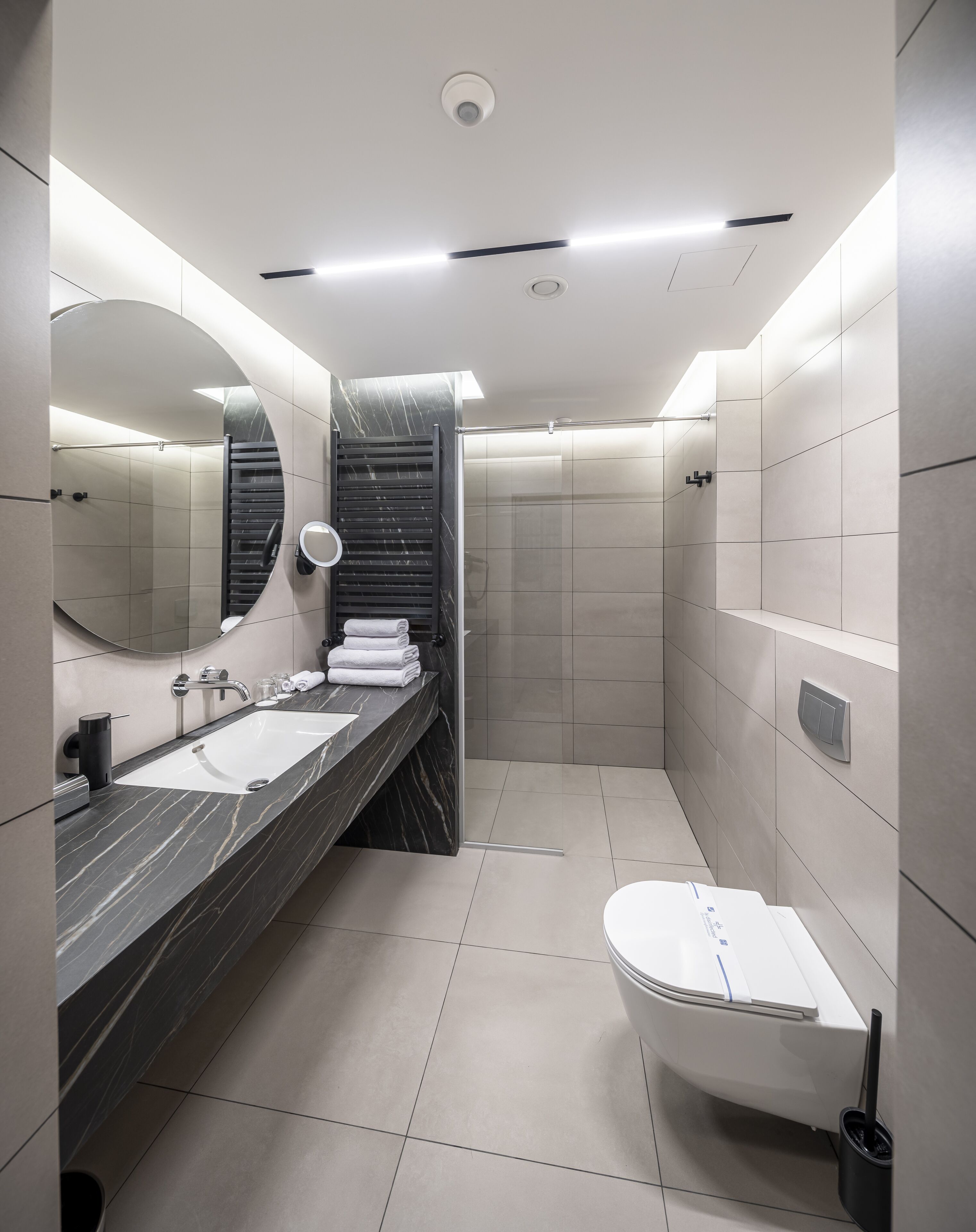 A modern bathroom with a dual sink vanity, a large mirror, a heated towel rail, and a walk-in shower.