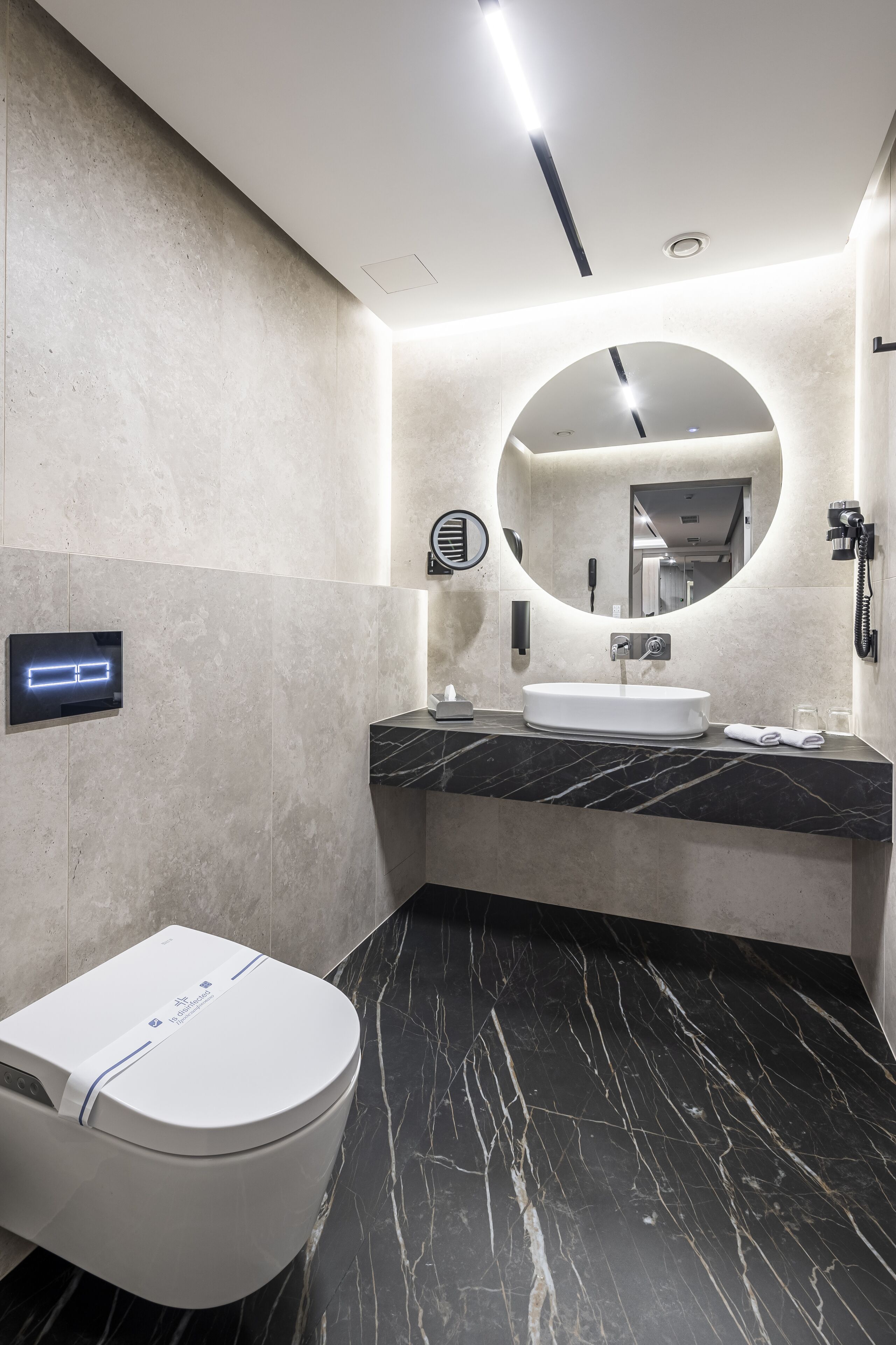 A modern bathroom with a marble floor, a white toilet, a sink with a mirror, and a digital clock.