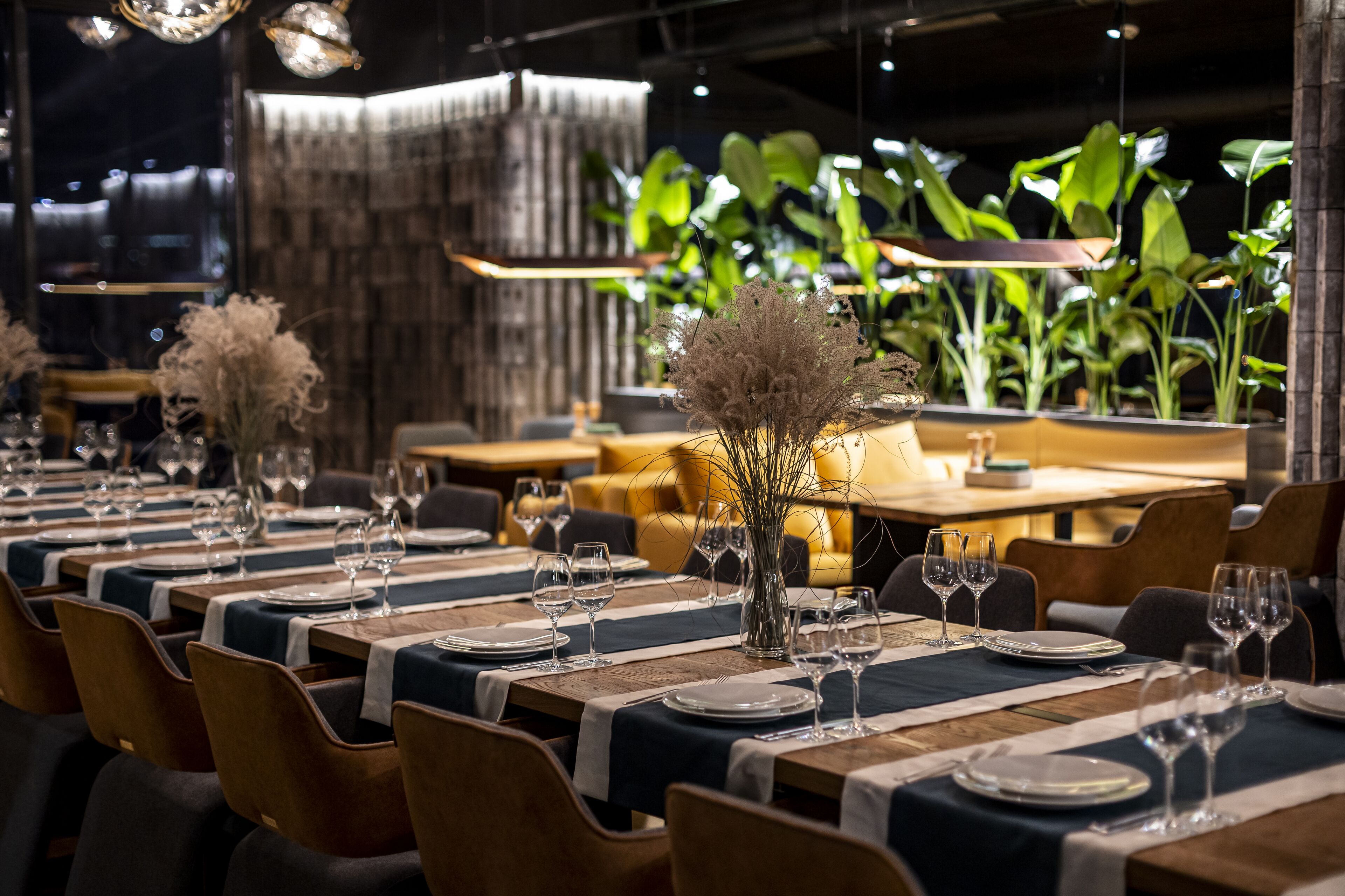 Un restaurante con largas mesas preparadas para comer, con un diseño moderno, sillas de madera y un centro de mesa decorativo con plantas.