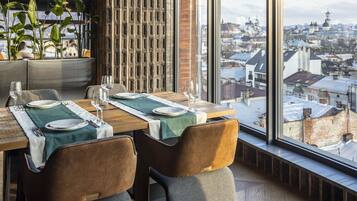 A dining area with a view of a cityscape through large windows, featuring a wooden table set with plates, glasses, and a green table runner.