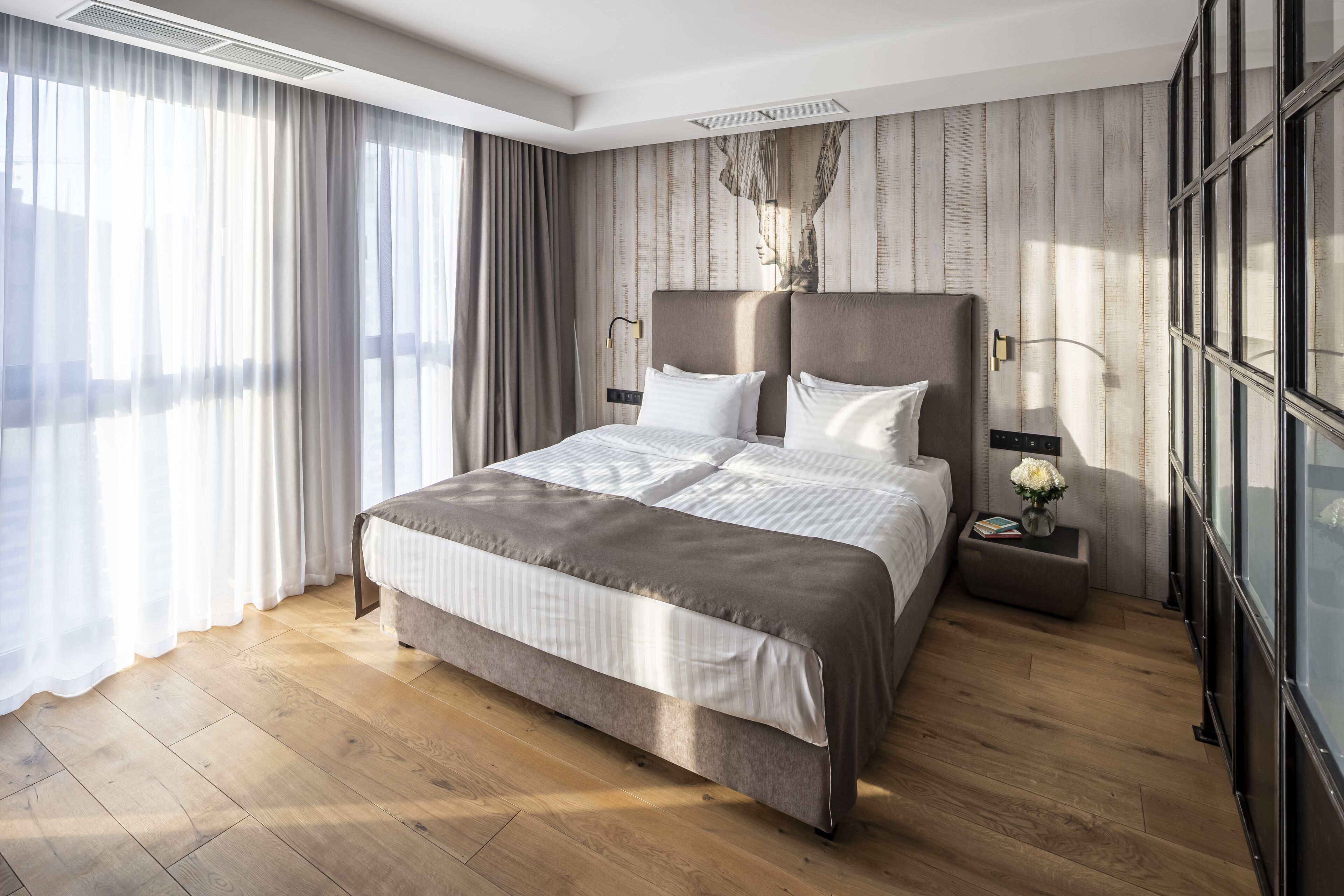 A modern hotel bedroom with a large bed, a nightstand, a wall-mounted lamp, and a large window with curtains.
