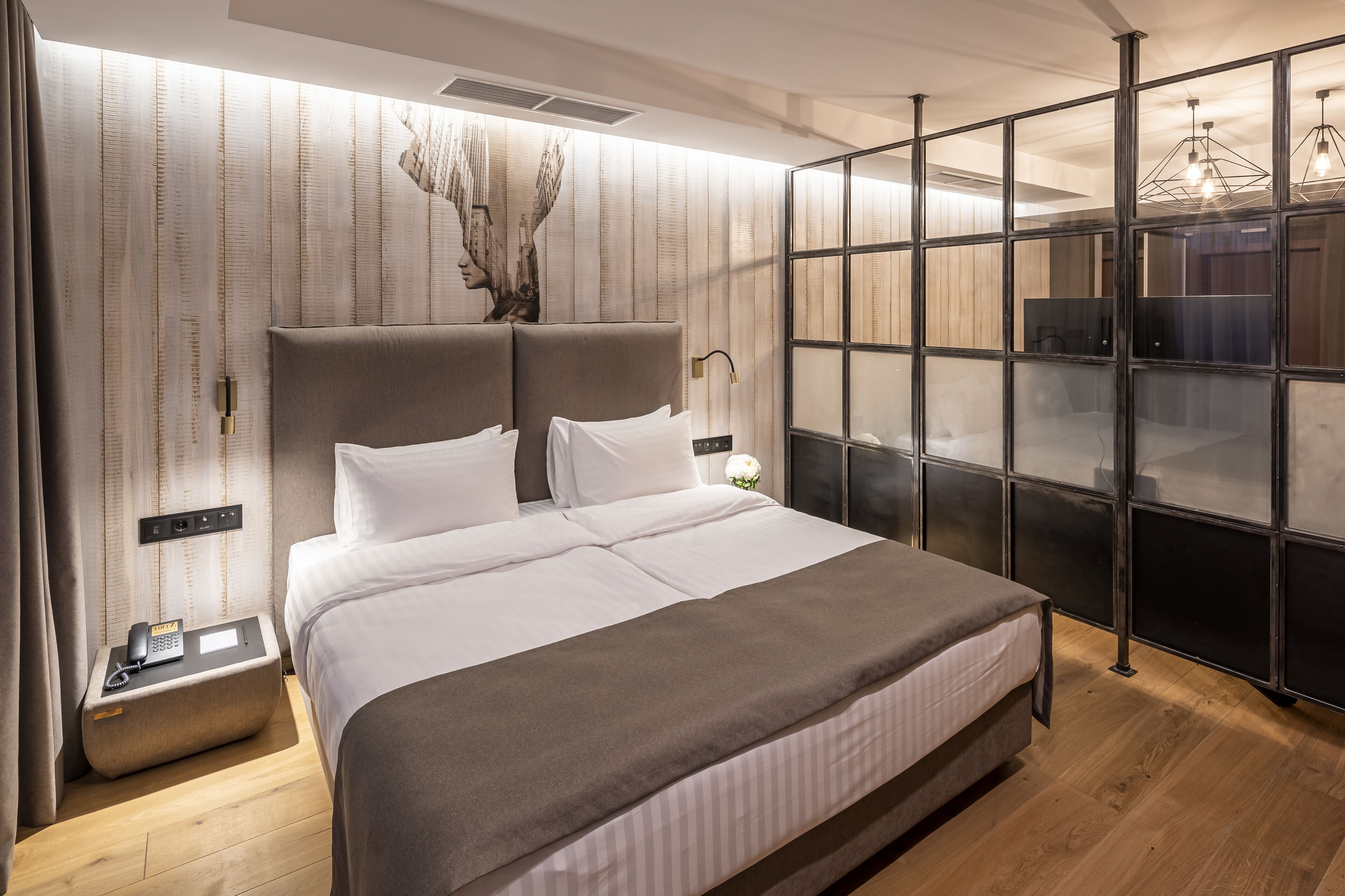 A modern hotel room with a large bed, a bedside table with a phone, a decorative wall piece, and a large window with a view.